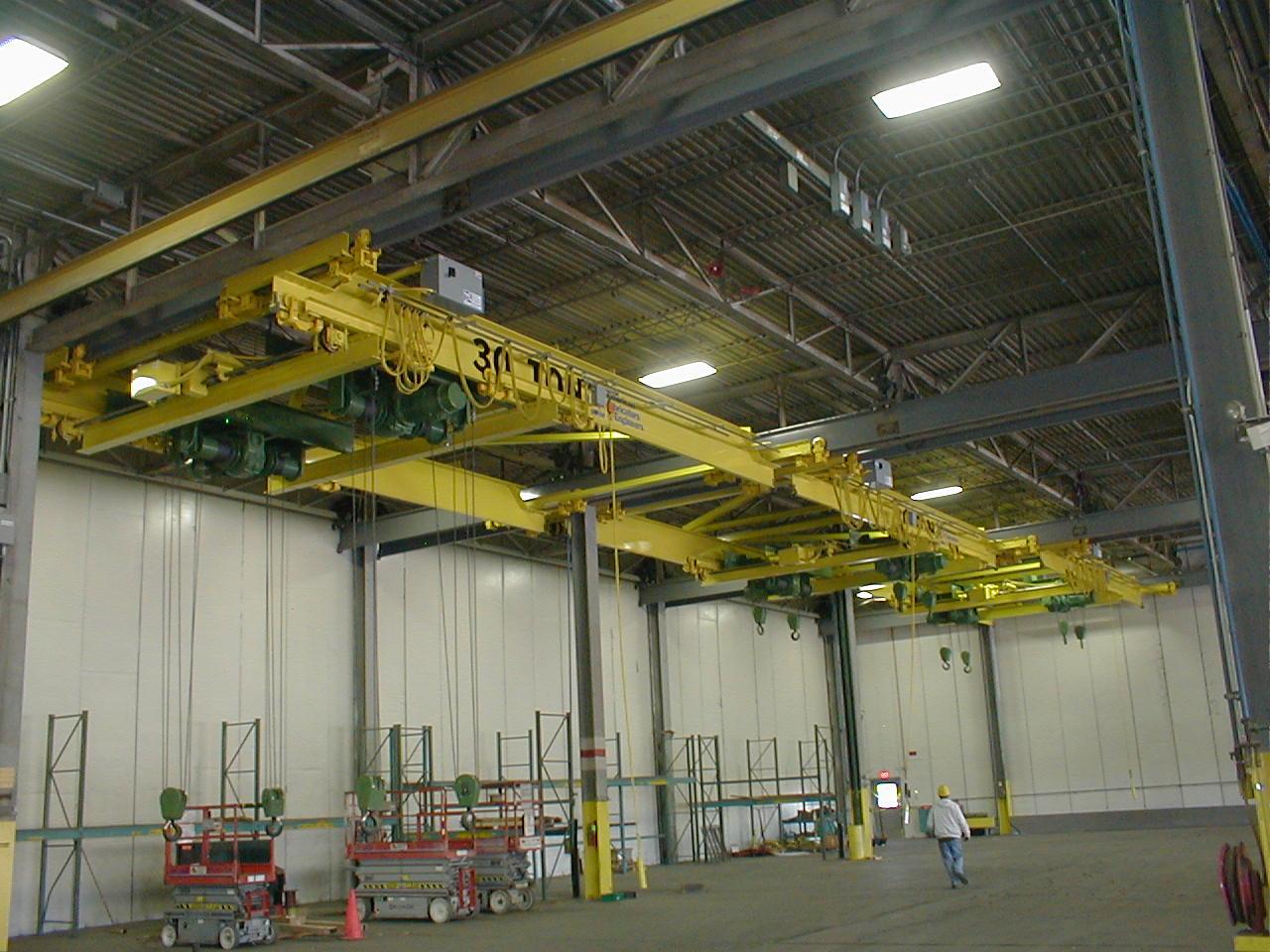 AFE Crane 30-ton under hung double girder patented track bridge crane system installed in a manufacturing facility. Each bridge crane is equipped with two dual-hook hoists, allowing synchronized lifting of heavy or oversized loads. The cranes are suspended from a patented track system supported by the building structure. Below, several scissor lifts and a worker in a hard hat suggest active setup or inspection.